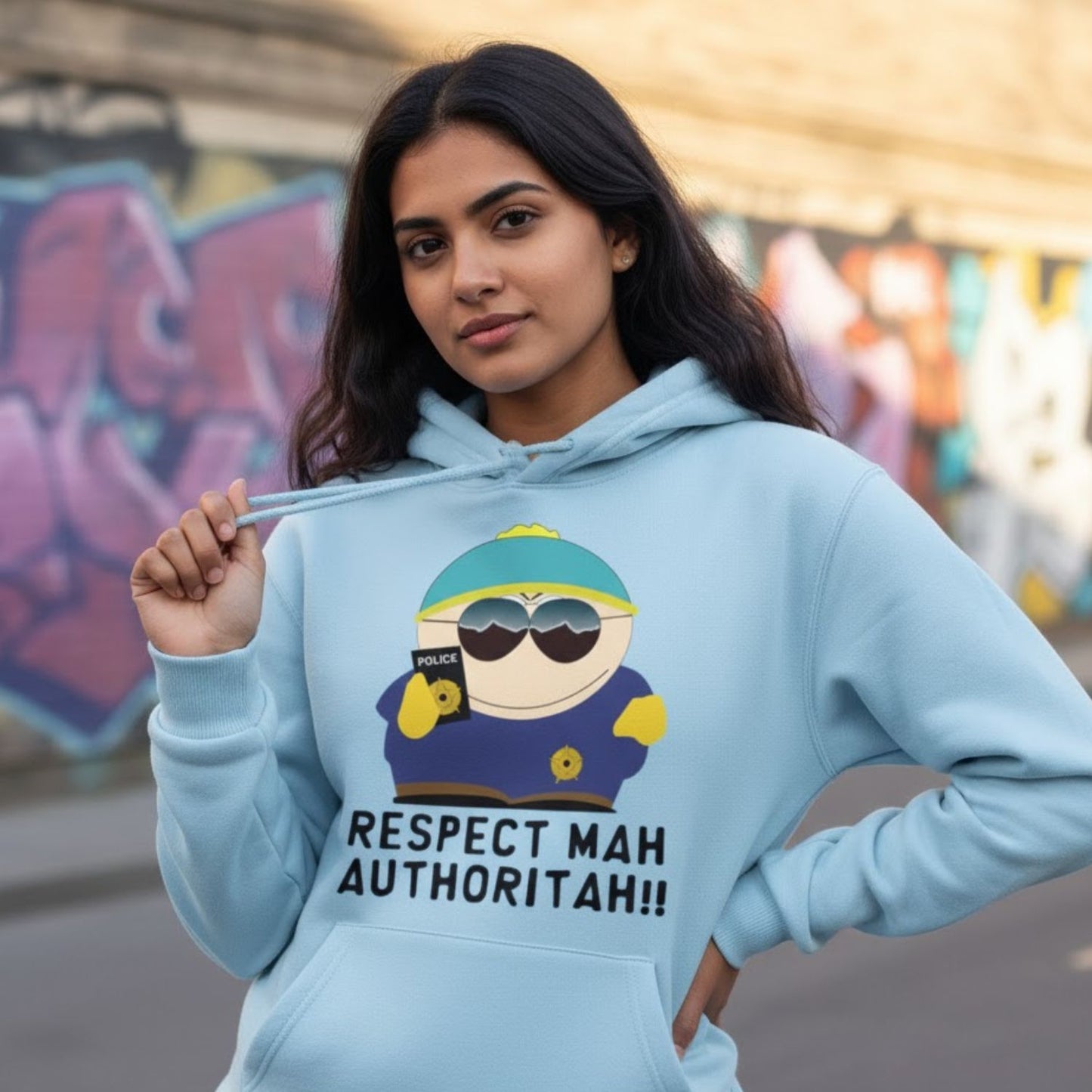 Person wearing a light blue hoodie with a graphic design and text, standing in front of graffiti. South Park Respect Mah Authoritah Hoodie by The Banyan Tee