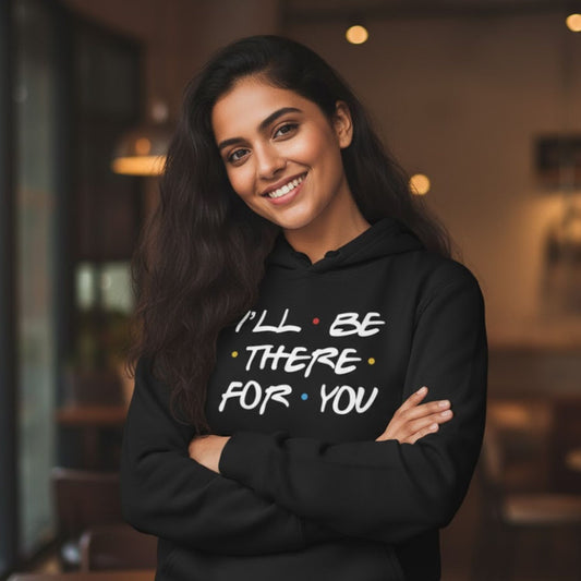 Woman wearing a black hoodie with 'I'll be there for you' text in a blurred indoor setting. Friends Hoodie. The Banyan Tee