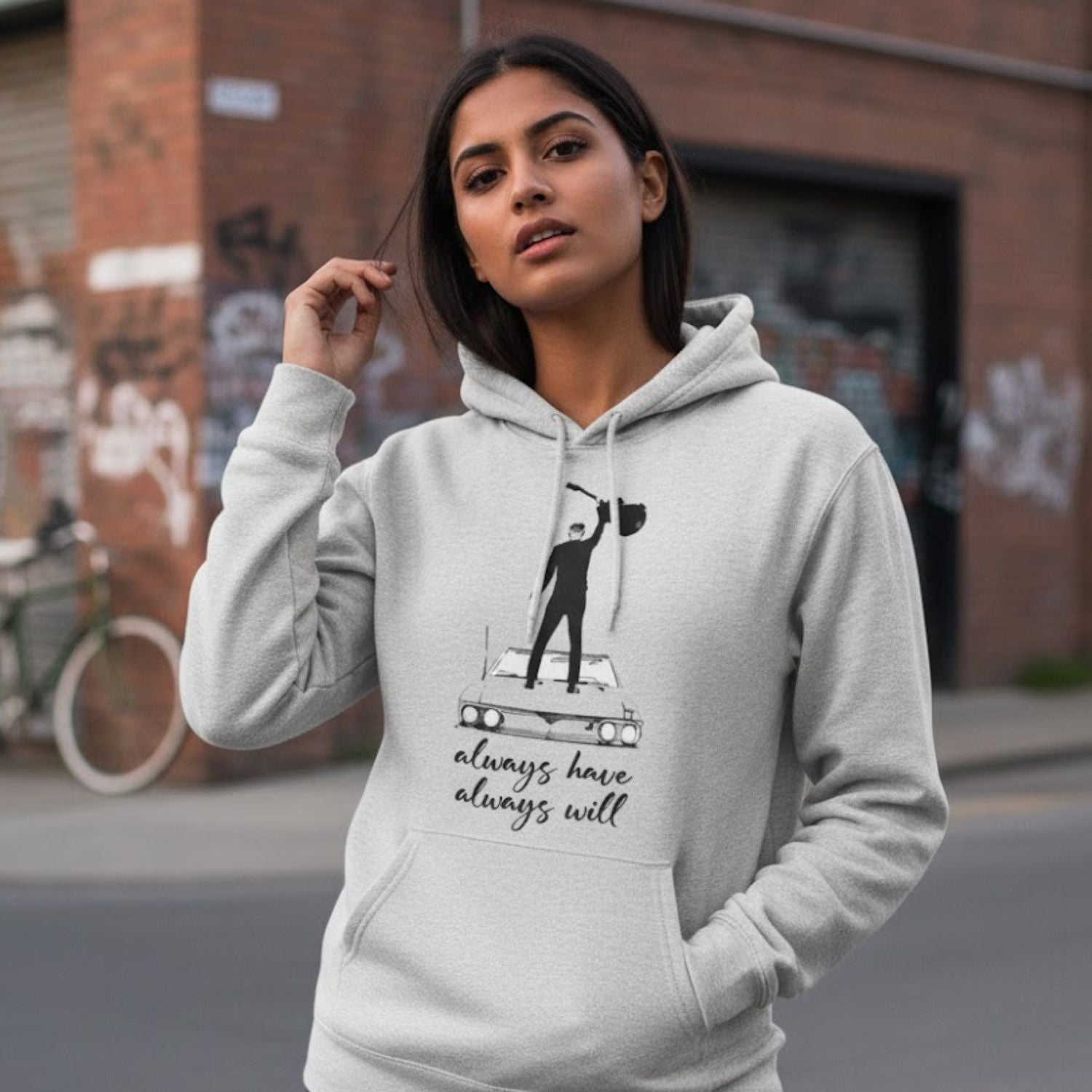 Woman wearing a gray hoodie with a graphic design and text, standing on a street. The Banyan Tee Bryan Adams Always Hav Always Will Hoodie.
