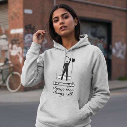 Woman wearing a gray hoodie with a graphic design and text, standing on a street. The Banyan Tee Bryan Adams Always Hav Always Will Hoodie.