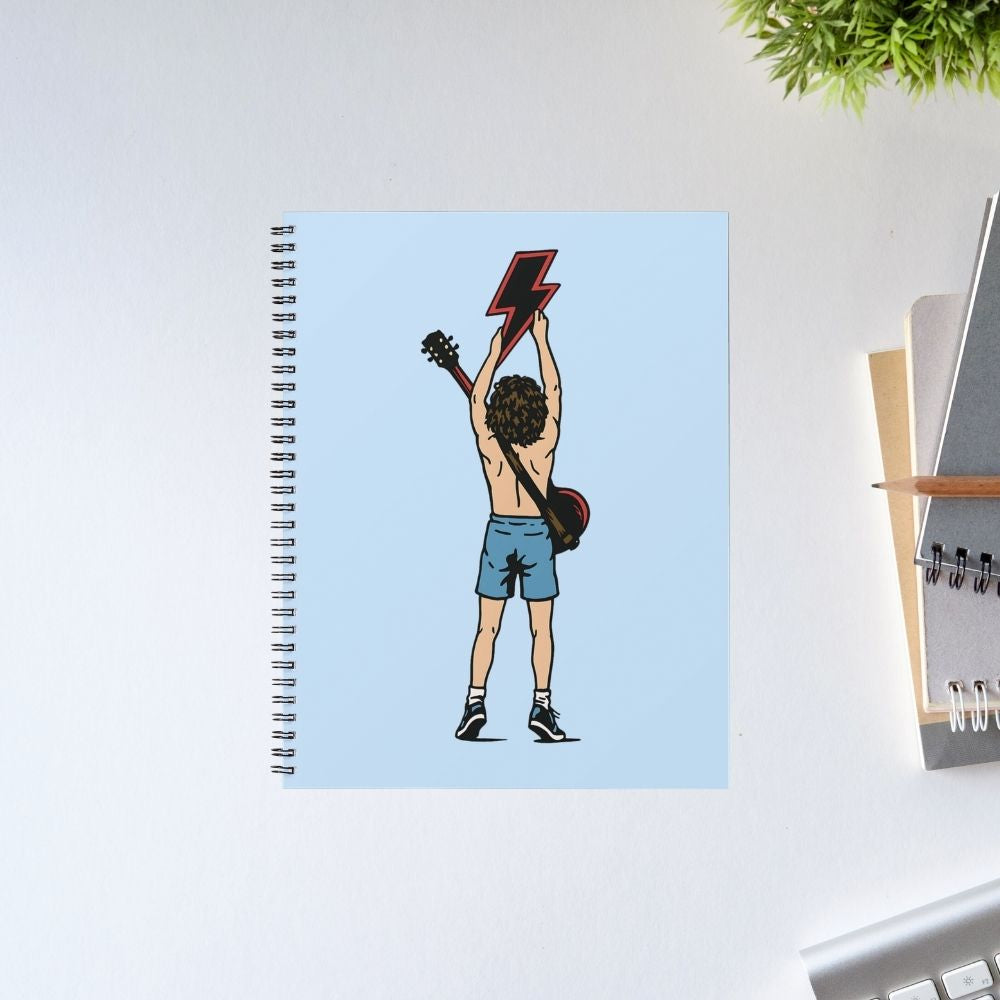 Notebook with a cover featuring an illustration of a person holding a guitar on a light blue background, placed on a desk. Notebook AC/DC The Banyan Tee