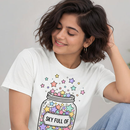 A woman wearing a cream-colored graphic t-shirt with a colorful star jar design and blue jeans, sitting in a bright indoor setting.