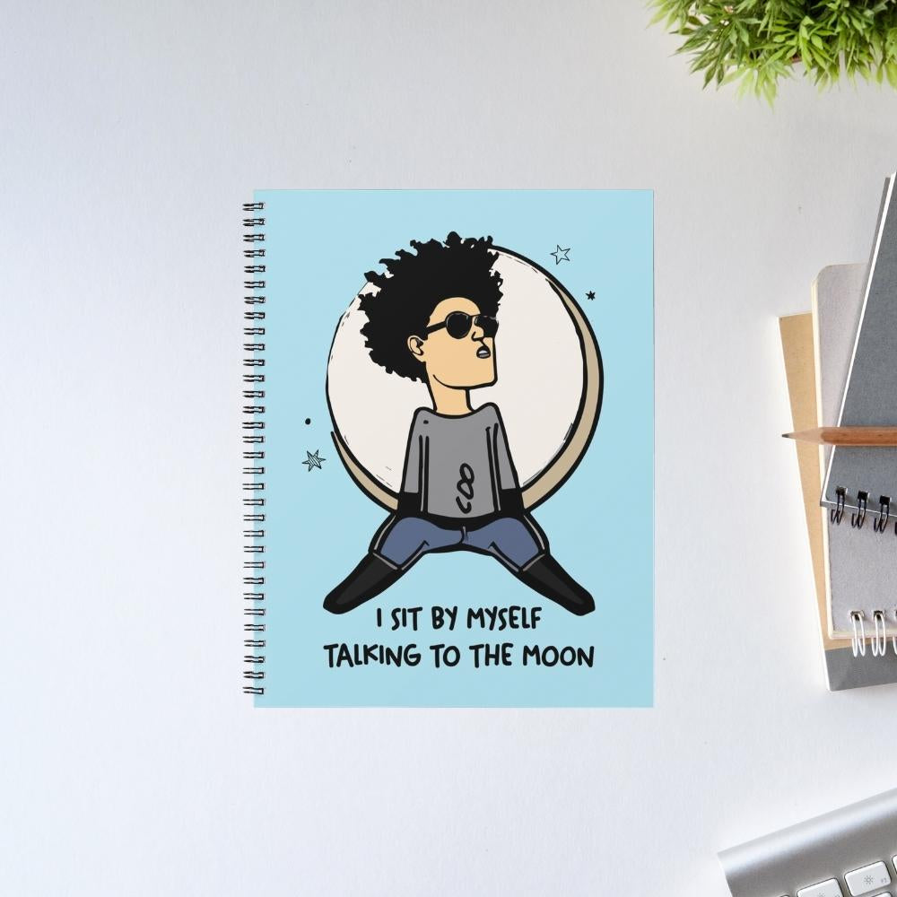 Notebook with a design of a person sitting on the moon and text 'I sit by myself talking to the moon' on a light blue background. Bruno Marsh Gift The Banyan Tee India