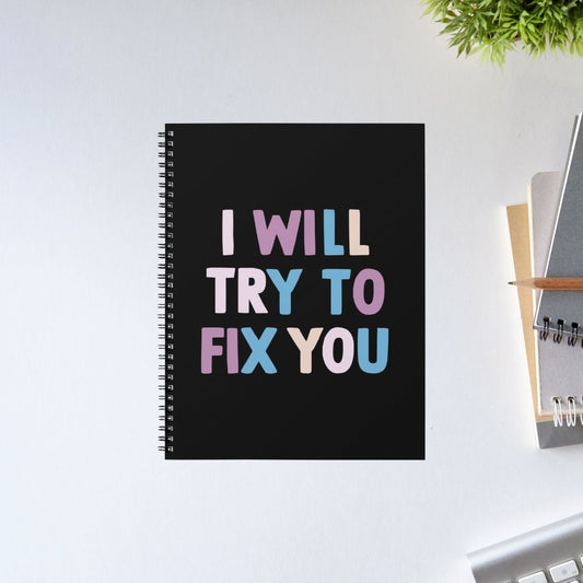 Black notebook with colorful text on a white surface with office supplies. Coldplay The Banyan Tee I Will Try To Fix You.