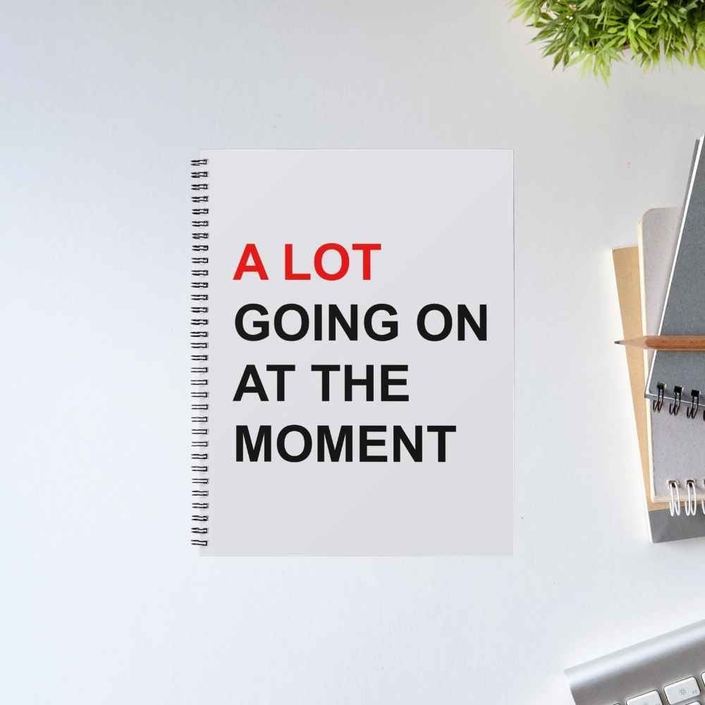 Taylor Swift A Lot Going On spiral notebook with bold red and black text 'A LOT going on at the moment' on a light grey cover – unruled notepad diary, The Banyan Tee