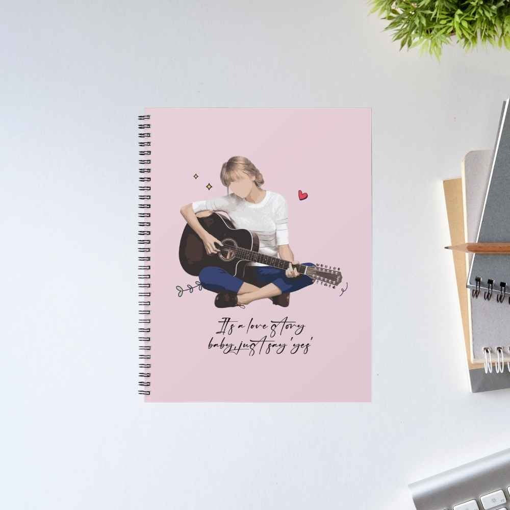 Taylor Swift Love Story spiral notebook with an illustrated guitarist and the lyric 'It's a love story, baby, just say yes' on a pink cover – unruled notepad diary, The Banyan Tee