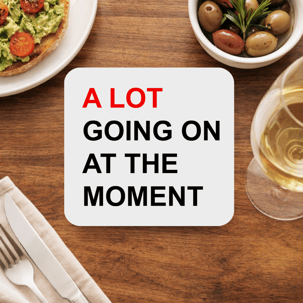 Taylor Swift A Lot Going On coaster in bold black block type with A LOT in red, on a light grey square beside avocado toast, olives and white wine